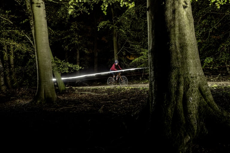 Lone rider at night on a woodland trail between large tree trunks, a streak of light trailing behind.