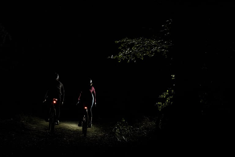 Rear view of two night riders on a dark woodland track, each bike carrying a glowing red tail light.
