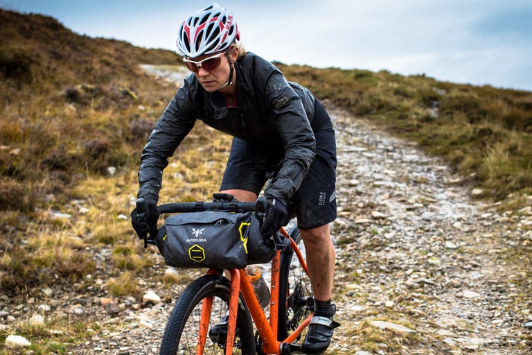 Naomi Freireich climbs a stony track in GORE Wear kit with an Apidura handlebar bag on her orange Mason Bokeh.