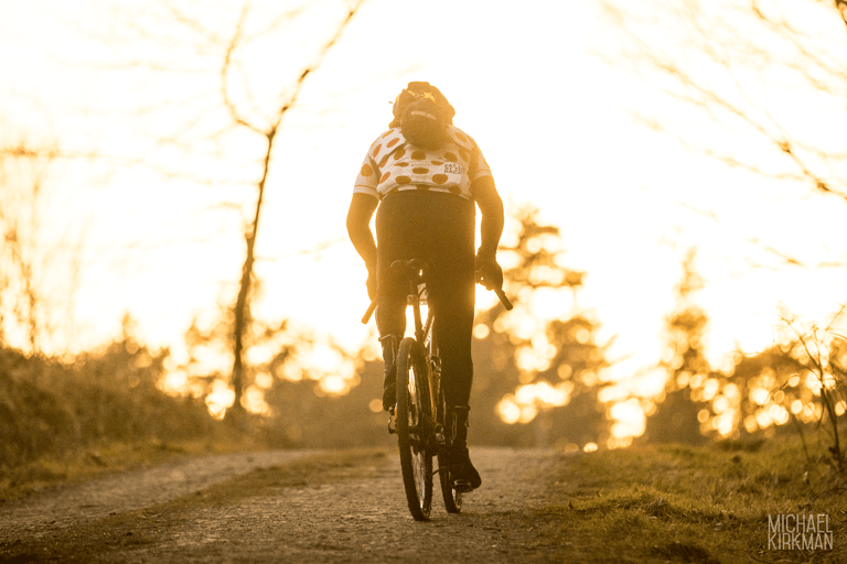 Rider in a polka-dot jersey heads up a gravel track into low evening sun at Yorkshire True Grit Dark Skies.