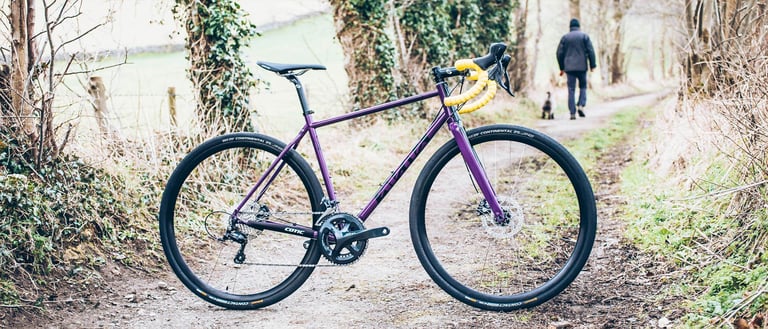 Purple Cotic Escapade with yellow bar tape and disc brakes parked on a green lane beside a field.