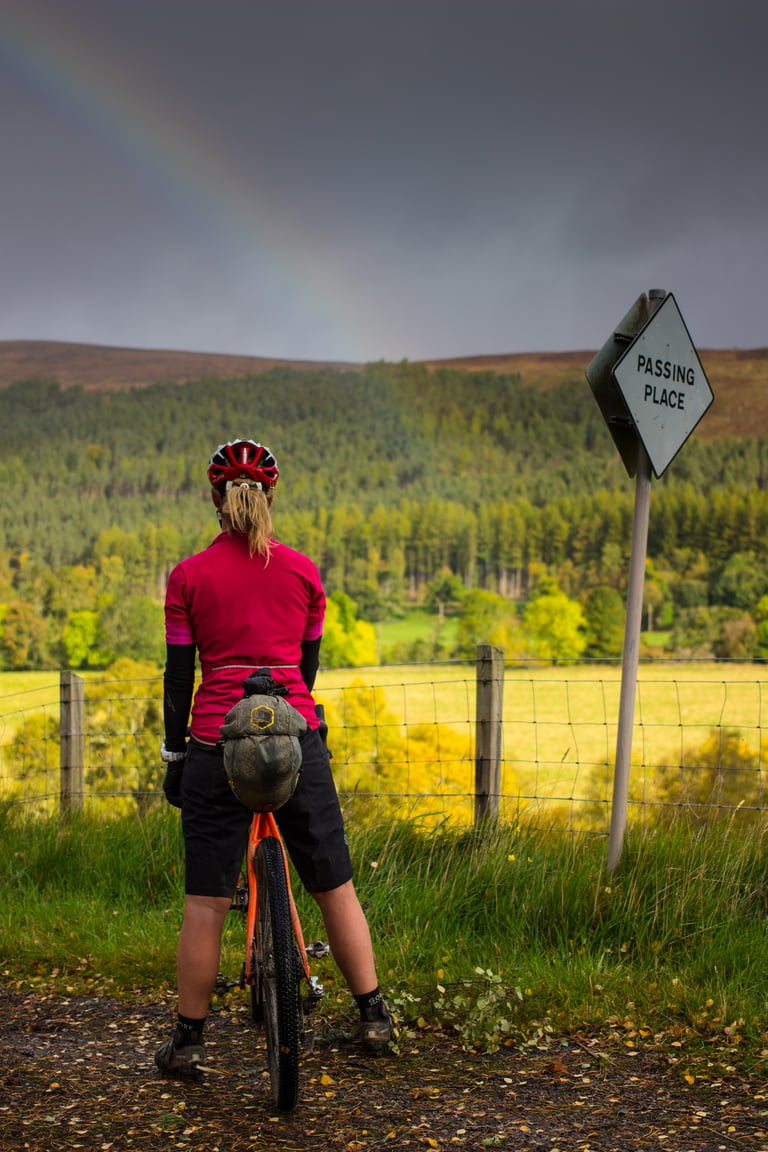 Naomi Freireich stands astride her bike watching a rainbow beside a Passing Place sign in the Scottish Highlands.