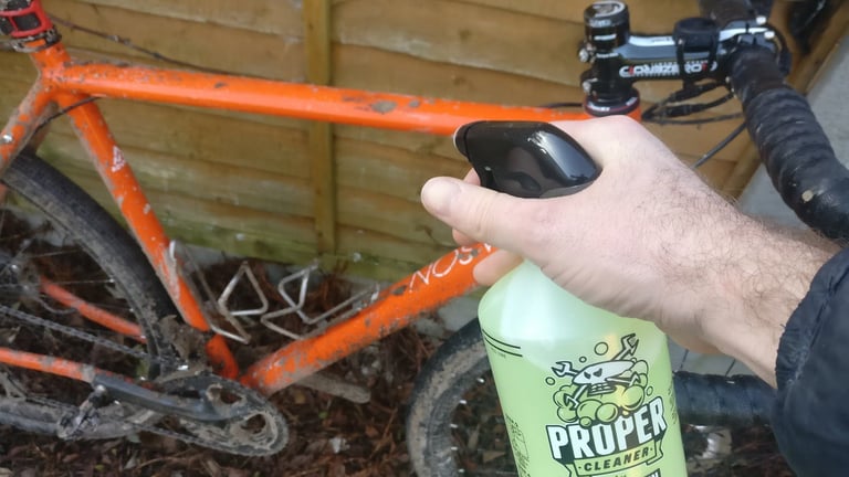 Hand holding a trigger bottle of Proper Cleaner, spraying a muddy orange gravel bike leaning against a garden fence.