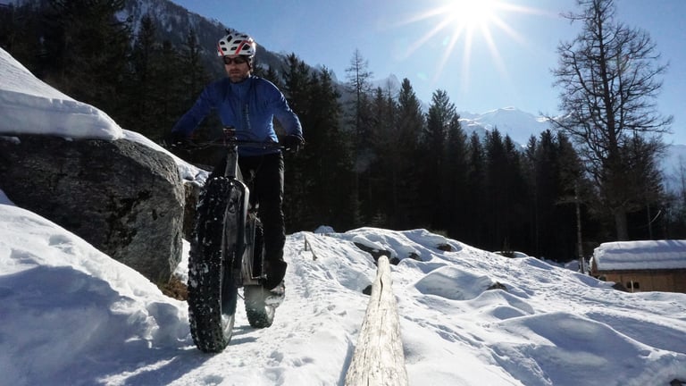 Rider on a Travers Bat Fastard fatbike crossing a snowy trail past a boulder and pine woods near Chamonix.
