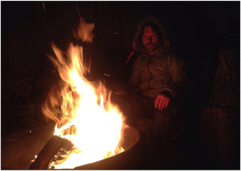 Simon Bryant sits beside a campfire at night in a hooded down jacket and head torch.