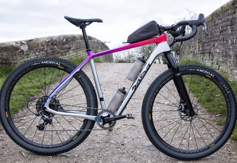 Salsa Cutthroat with a pink-to-silver fade frame and top-tube bag, side view on a track beside a stone bridge.