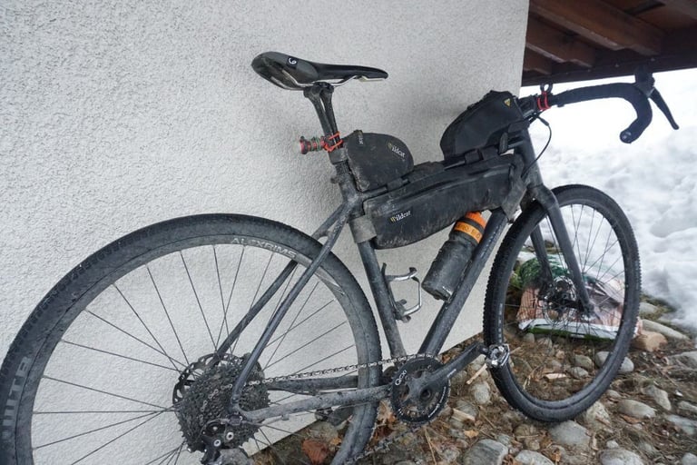 Muddy Pinnacle Arkose with Wolftooth Tanpan 1x drivetrain and frame bags, leaning against a wall beside snow.