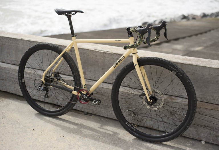 Quicksand Fearless Vulture with camouflage bar tape and a single-ring crankset leaning on a timber sea wall.