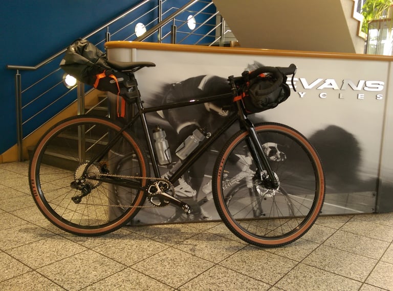 Black drop-bar adventure bike with bikepacking seat pack and bar bag parked against an Evans Cycles display.