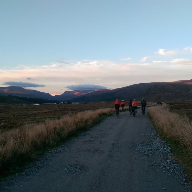 Group of riders in red and orange jackets head along a gravel track at dusk towards hills and a stretch of water.