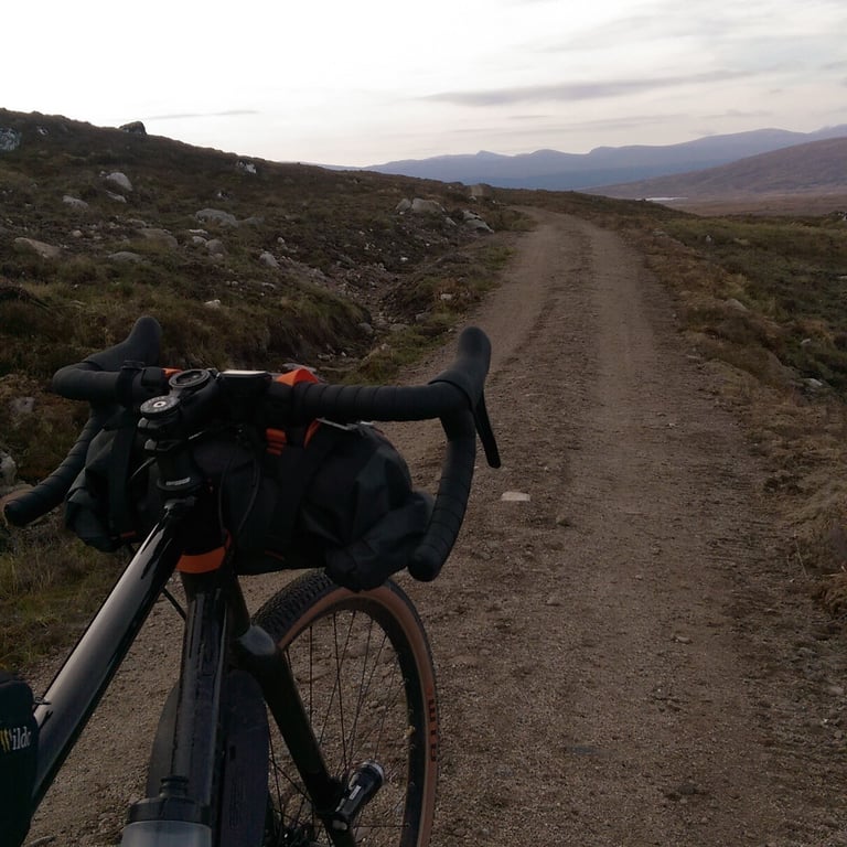 View over a gravel bike's drop bars and bar bag down a stony moorland track towards distant hills.