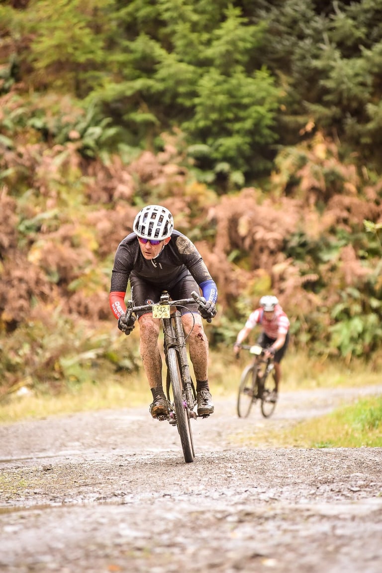 Mud-spattered rider numbered 142 races a black gravel bike up a forest track, with a second rider behind.
