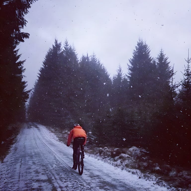 Rider in an orange jacket on the Salsa Cutthroat pedals a snow-covered forest road through falling snow in Kielder.