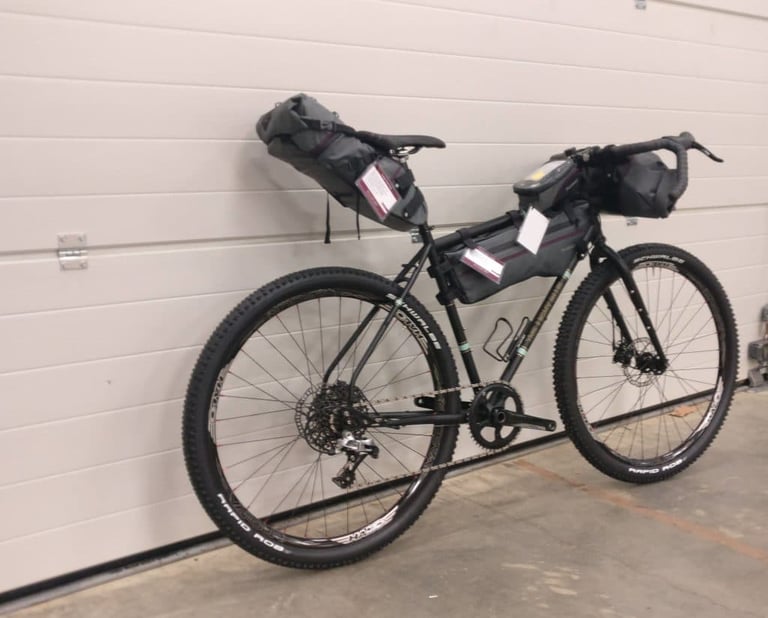 Light Blue Darwin One By MT with Passport bikepacking luggage, rear three-quarter view against a garage door.