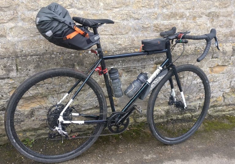All-City Macho Man in audax trim with an Ortlieb seat pack, top-tube bag and two bottles against a stone wall.