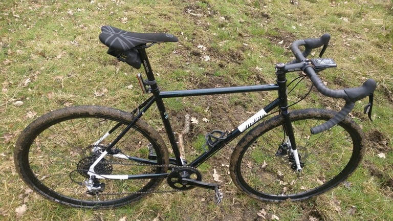 All-City Macho Man with mud-caked tyres lying on a grassy, churned-up trail.