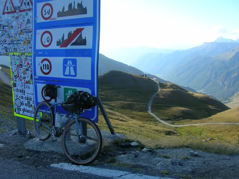 Loaded bike leaning on sticker-covered road signs at an alpine pass on the Turin Nice Rally, gravel track winding below.