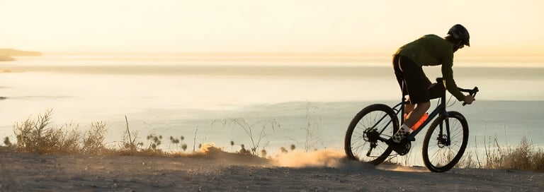 Rider on a Salsa Journeyman kicks up dust on a dirt track above calm water in hazy low light.