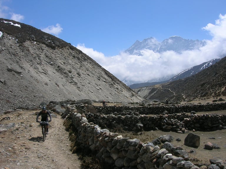 Mountain biker rides a dirt track beside a dry-stone wall below snowy Himalayan peaks in Nepal.