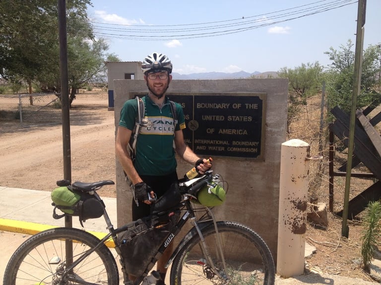 James Olsen in an Evans Cycles jersey with a loaded mountain bike at the US boundary marker after finishing the Tour Divide.