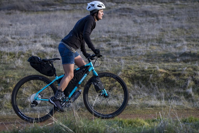 Rider on a loaded Surly Bridge Club with frame and seat bags on a dirt track across open grassland.