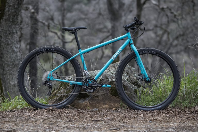 Surly Bridge Club in Diving Board Blue with flat bars and WTB 650b tyres, parked against a boulder in woodland.