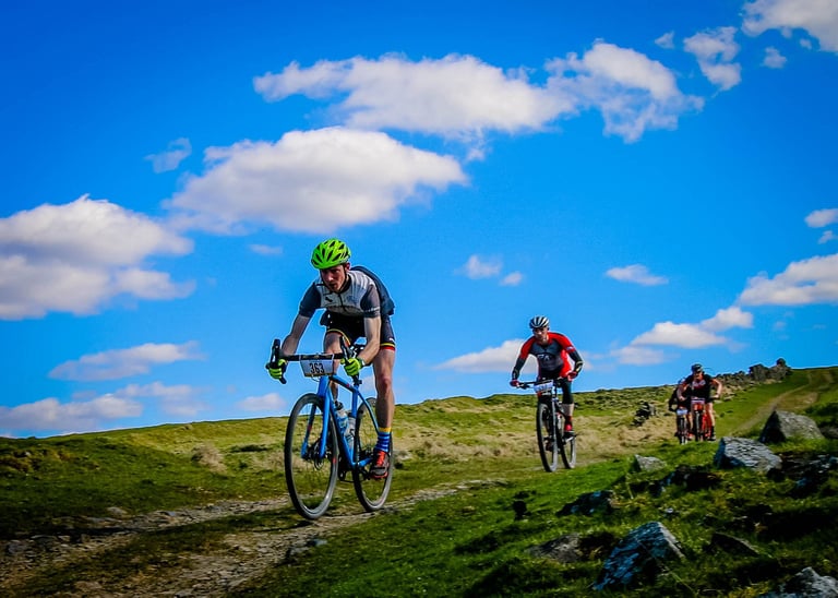 Rider in a green helmet on a blue gravel bike leads a group down the grassy Jimmy's Gate descent under blue skies.