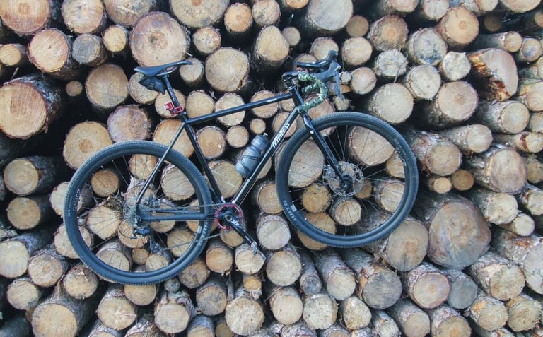 Fearless Vulture gravel bike with bottle and saddle bag propped against a tall stack of cut logs.