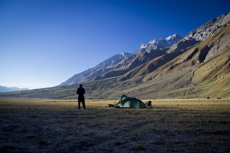 Green tent and a standing figure at a camp on open ground below snow-capped peaks, from Escape By Bike.