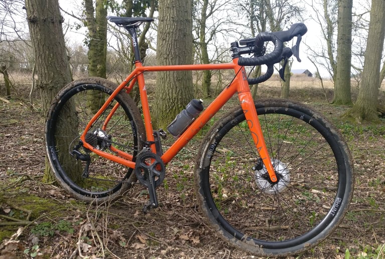 Orange Mason Bokeh gravel bike fitted with Hunt 30 Carbon Gravel wheels, parked among woodland trees.