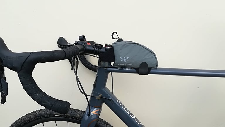 Apidura top tube bag strapped behind the stem of a grey Mason Bokeh gravel bike, seen side-on against a cream wall.