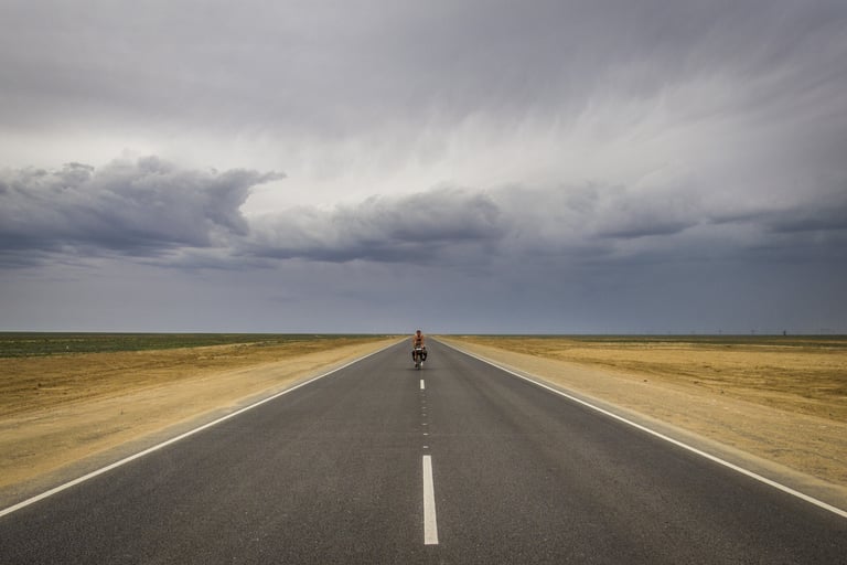 Lone loaded touring cyclist on a straight empty road under a heavy grey sky, from Joshua Cunningham's Escape By Bike.