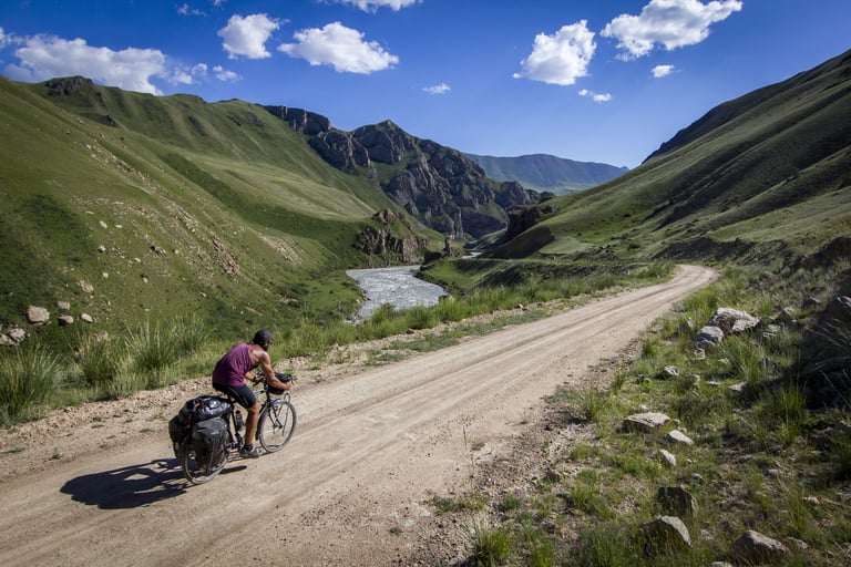 Cyclist with panniers rides a dirt road above a river in a green mountain valley on the Escape By Bike journey.