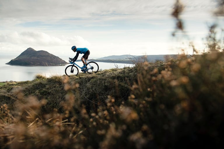 Grinduro rider on a blue gravel bike rides a grassy ridge above the sea on the Isle of Arran.