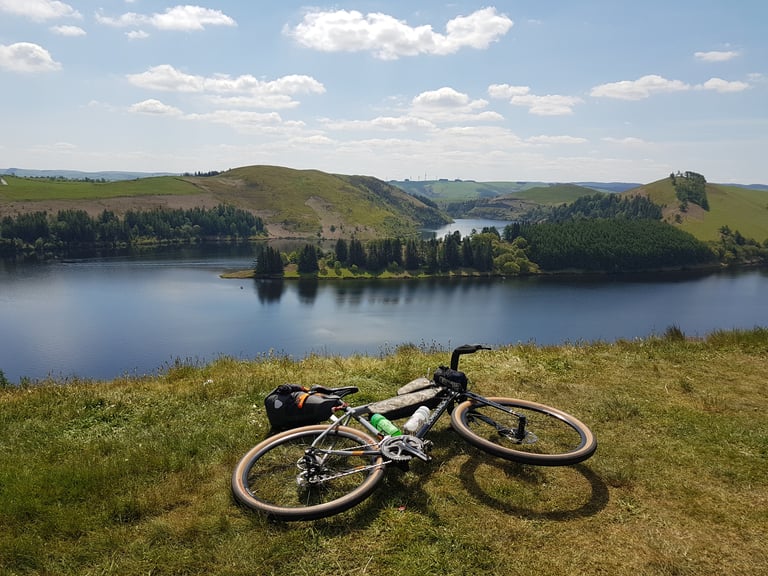 Loaded Bombtrack Audax lying on grass above a reservoir with forested hills beyond.