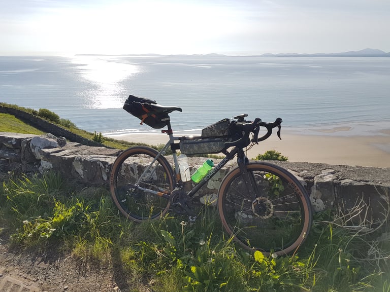 Loaded Bombtrack Audax with saddle and frame bags on a stone wall above a beach, hills across the bay.
