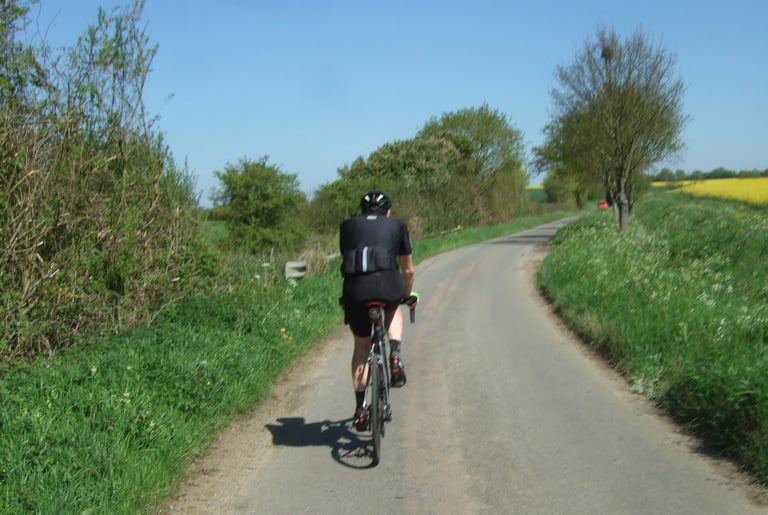 Rider in the black Le Col HC Clima jersey cycles away along a quiet lane, with a yellow rapeseed field beyond.