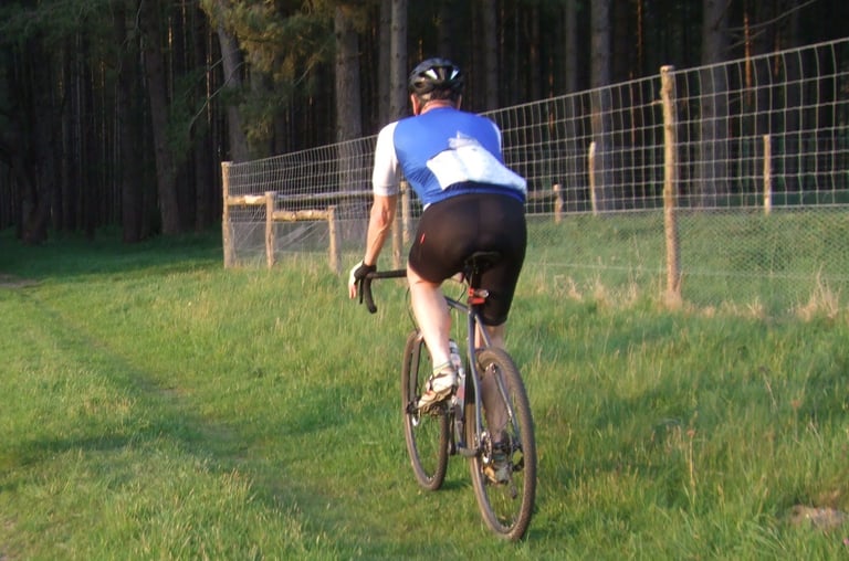 Rider wearing the Galibier Map Jersey cycles away along a grassy track beside a fence, with woods behind.