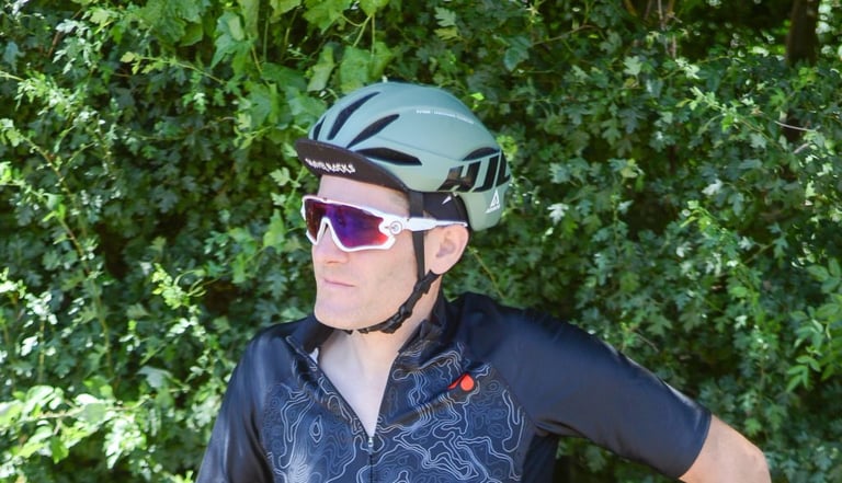 Rider wearing the green HJC Furion helmet with a cycling cap underneath and sunglasses, in front of a hedge.