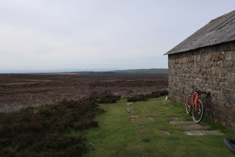 Orange gravel bike leaning against a stone shooting house on open heather moor on the private Yorkshire True Grit estate.
