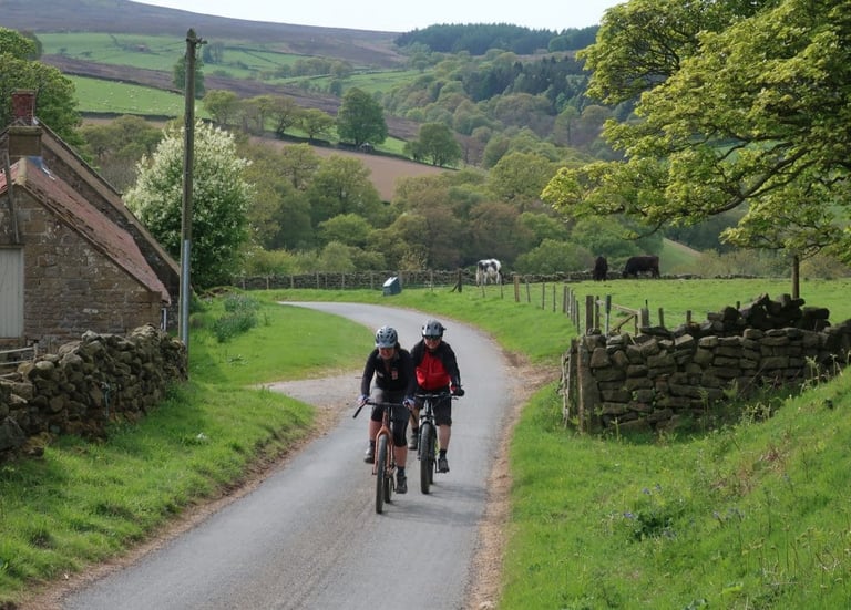 Debs and Andy ride up a narrow lane past stone walls and farm buildings towards the Yorkshire True Grit moorland trails.
