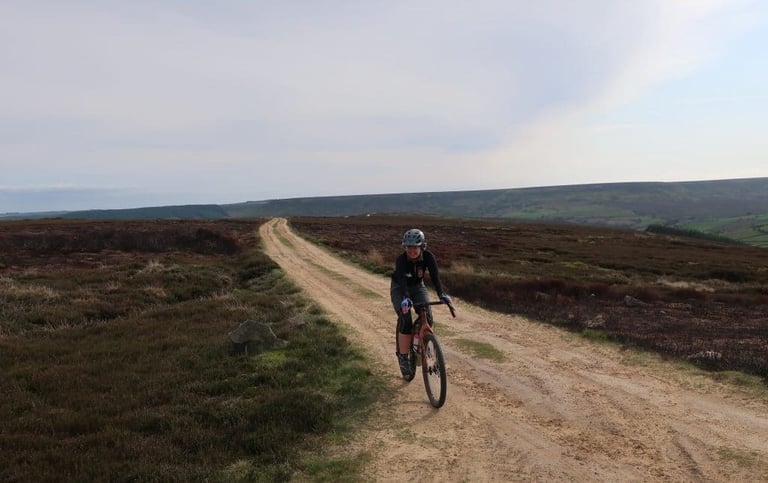 Rider on a gravel bike climbs a sandy estate track across heather moor on Whorlton Moor, North Yorkshire.