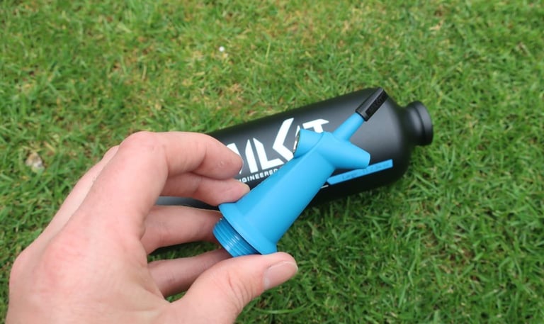Hand holding the blue plastic MilKit Booster head with its presta valve and threaded base, beside the bottle.