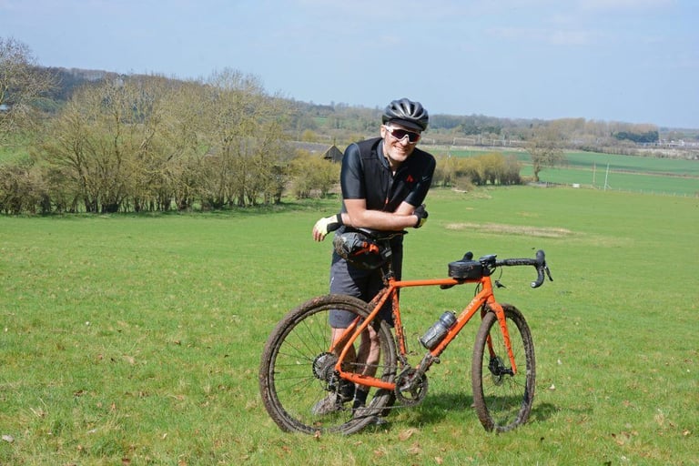 James Deane leans on his mud-spattered orange Mason Bokeh gravel bike in a green Chilterns field.