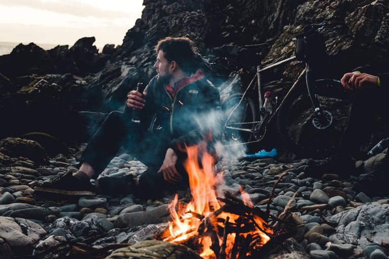 Rider with a bottle sits by a driftwood campfire on a pebble beach, a loaded bikepacking bike behind him.
