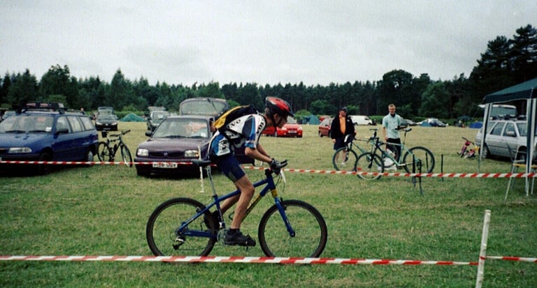 Young Josh Ibbett in a helmet races a mountain bike past taped course markers at a grassy cross-country race venue.
