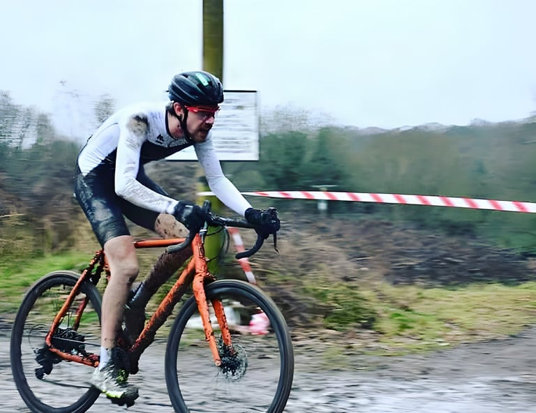 Mud-spattered Josh Ibbett on an orange Mason Bokeh races past course tape at a cyclocross event.