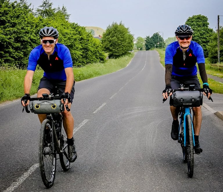 Two smiling riders with handlebar bags set off along a quiet road at the start of The Distance weekend.
