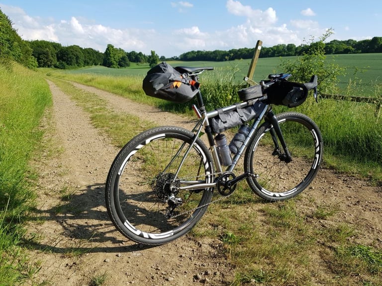 Reilly Gradient with Ortlieb bikepacking bags stands on a double-track farm lane between fields in summer sun.