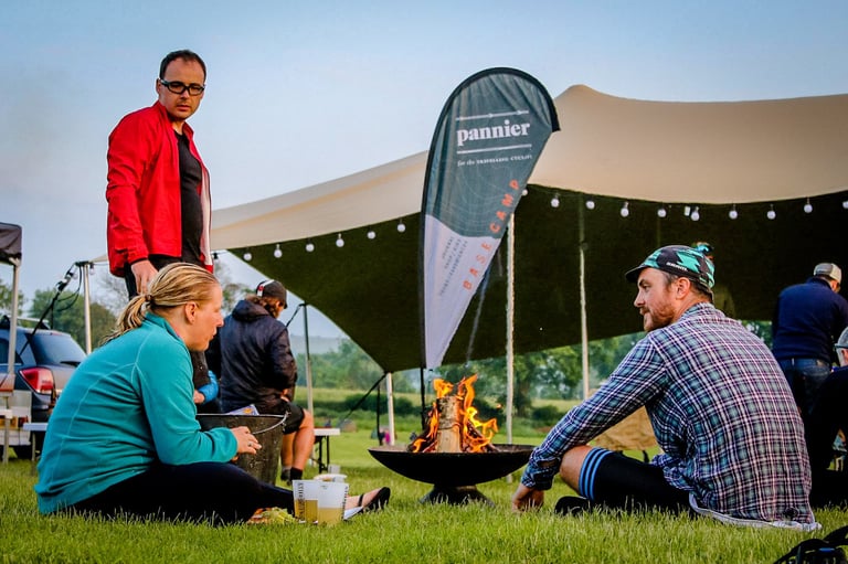 Riders sit round a fire pit beside the Pannier basecamp tent at The Distance overnight stop, Barn Farm Camping.